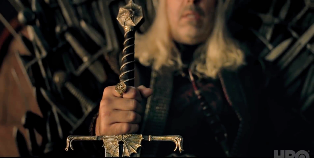 Paddy Considine's Viserys I Targaryen, sitting on the Iron Throne, grasps his sword as he grimaces; 